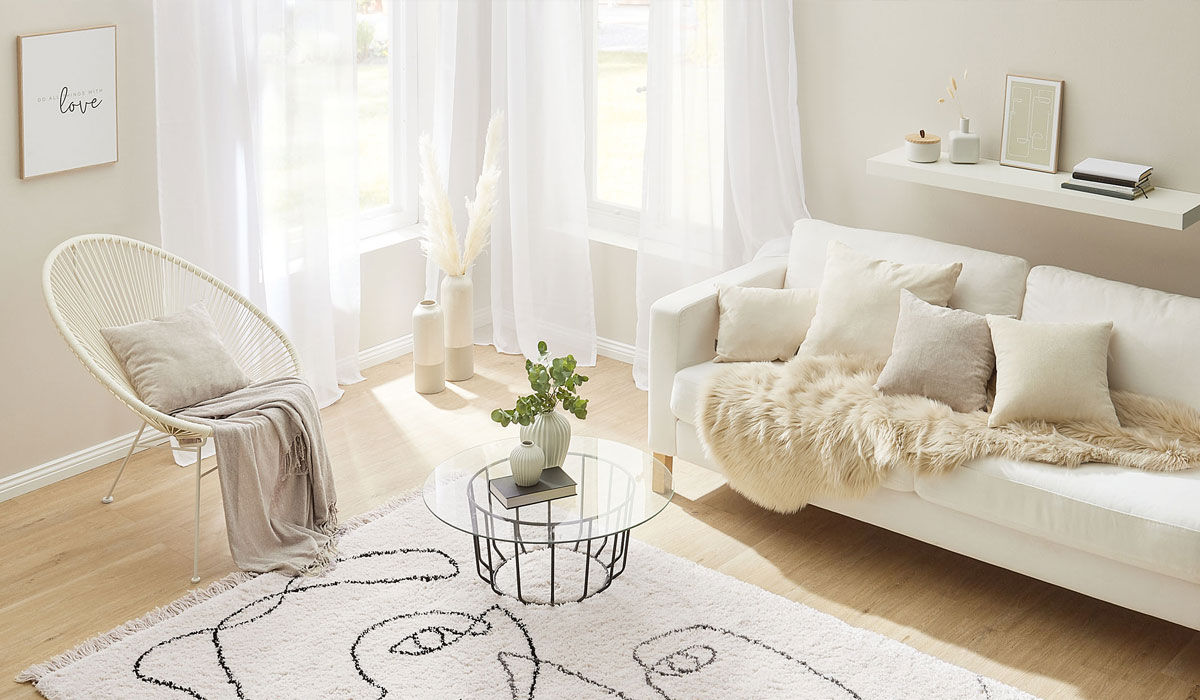 Sessel mit Decke und Kissen steht neben Glas-Couchtisch auf Teppich im hellen Wohnzimmer.