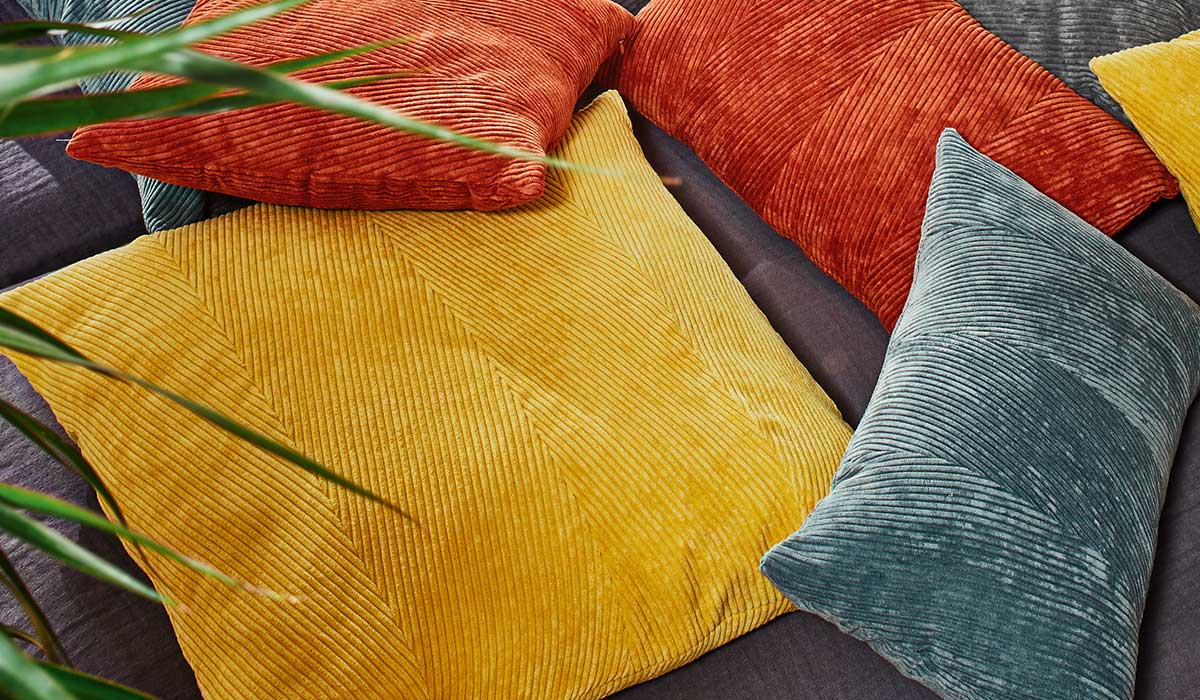 Mehrere Kissen in Gelb, Orange und Blau liegen auf einem dunklen Sofa mit grünen Pflanzenteilen.