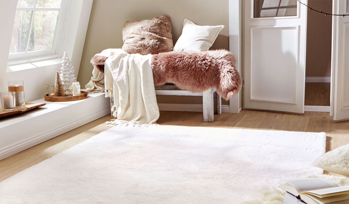 Bank liegt mit Decke und Kissen neben Fenster in hellem Zimmer mit Teppich auf Holzboden.