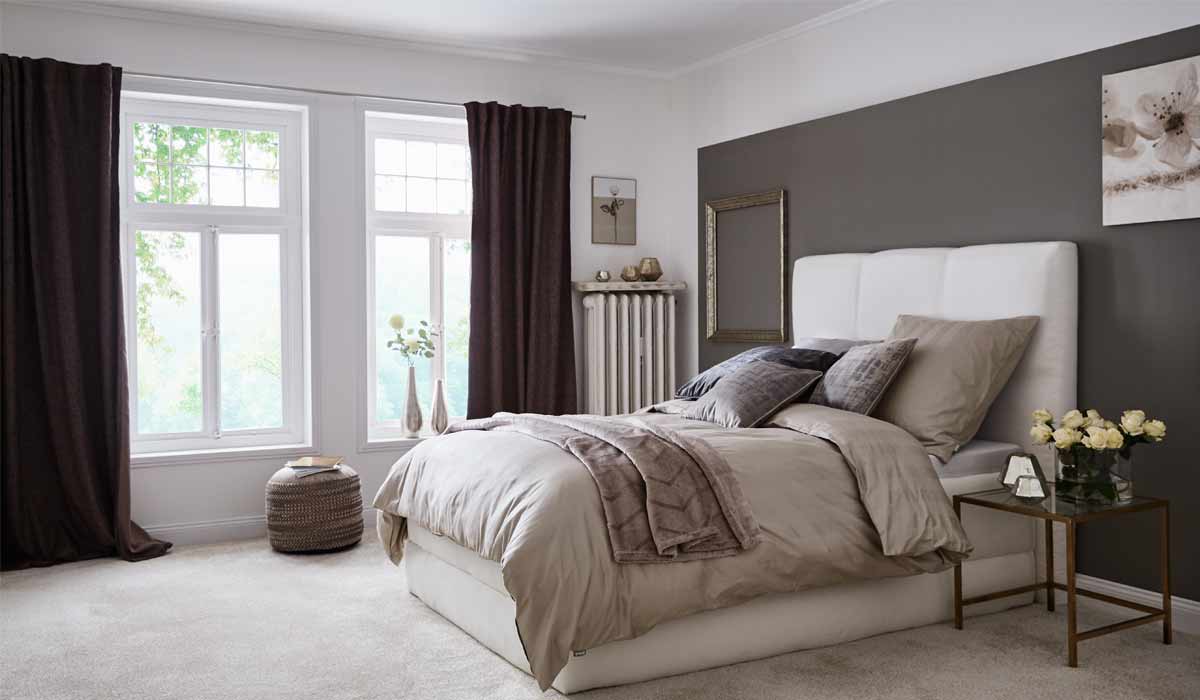 Bett liegt mit Kissen und Decke in modernem Schlafzimmer vor Fenster mit dunklen Vorhängen.